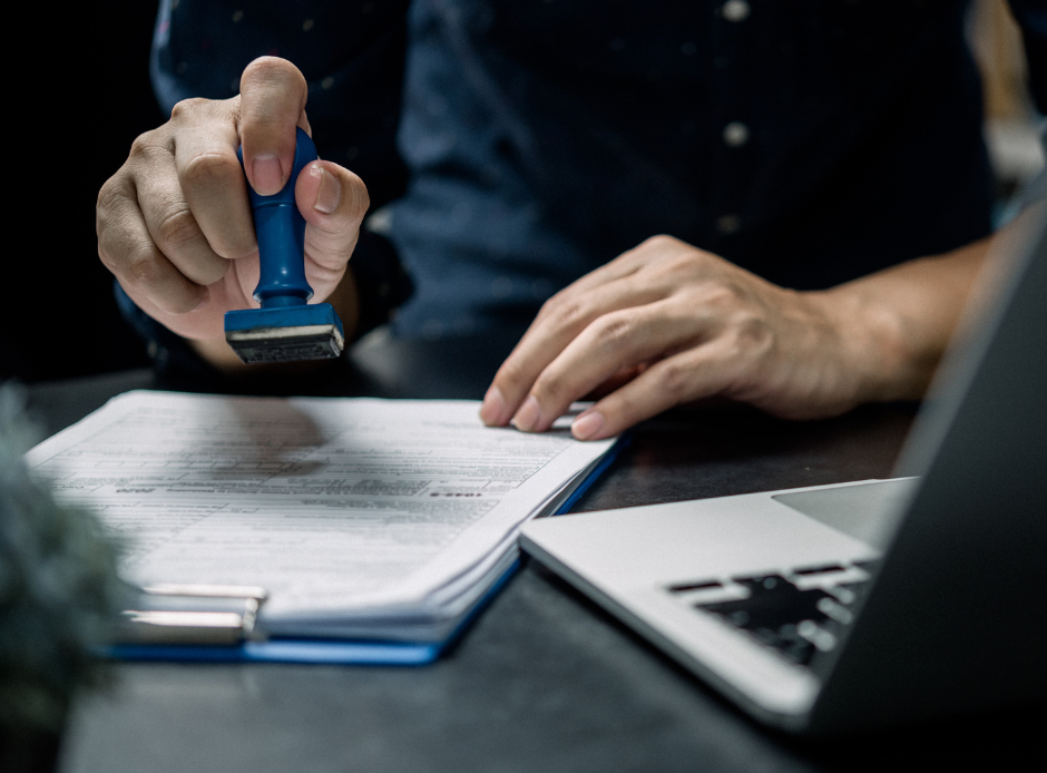 egy férfi a laptop mögött pecsétel egy papírt kék bélyegzővel